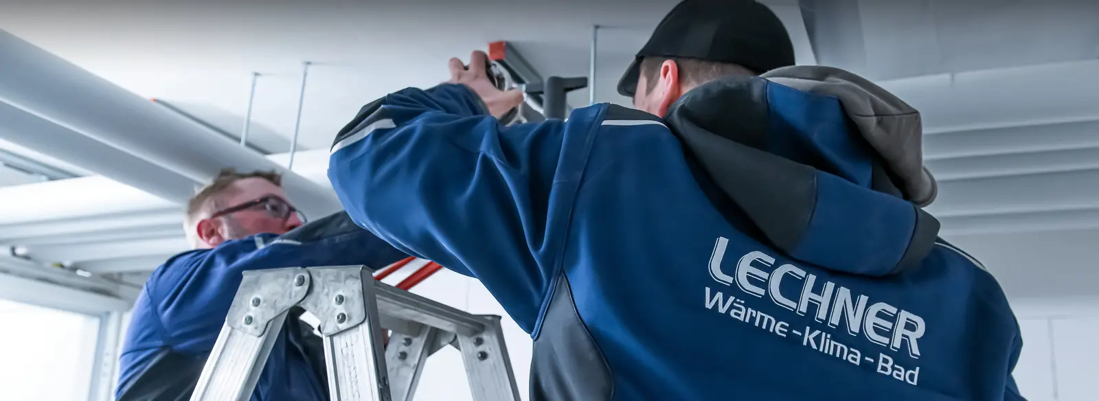 Das Bild zeigt zwei Männer in komfortabler, moderner DBL Arbeitskleidung, veredelt mit einem Rücken-Emblem „LECHNER Wärme-Klima-Bad". Der linke Mann steht mit dem Rücken zur Kamera, während der rechte konzentriert arbeitet, dabei zufrieden aussieht und ein Werkzeug hält.